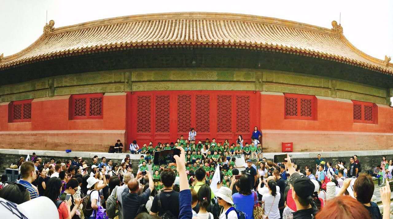 Group photo of elementary school students in front of Beijing's Forbidden City in front of an audience of their family members. 