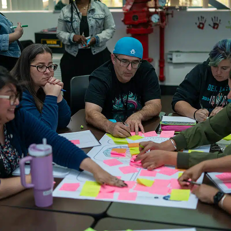 A group of people sit around a table covered with colorful sticky notes, collaborating and writing ideas. Some look thoughtful, while others reach for notes, suggesting a brainstorming session for planned experiments and improvement.