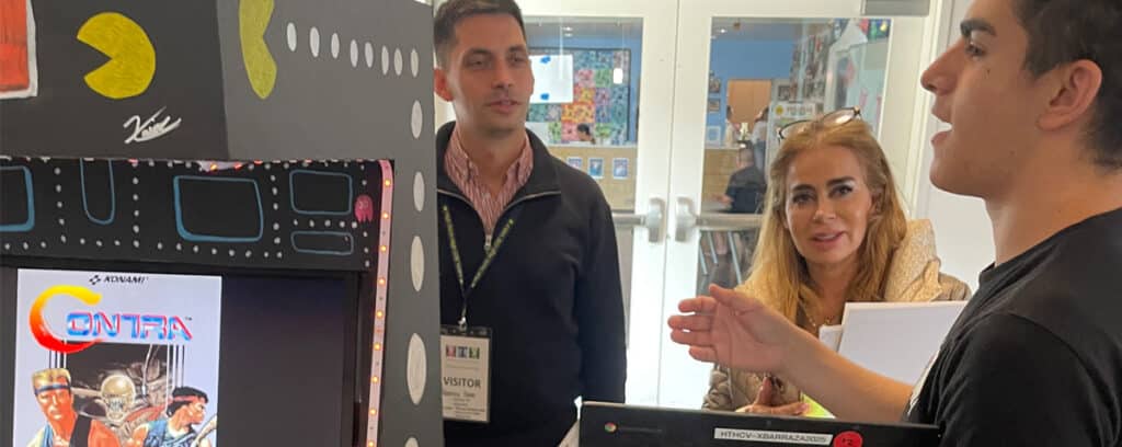 A young man explains an arcade game display to two visitors during one of the Curated Visits. The display features Pac-Man graphics and a screen showing the game Contra, capturing the guests attentive interest.