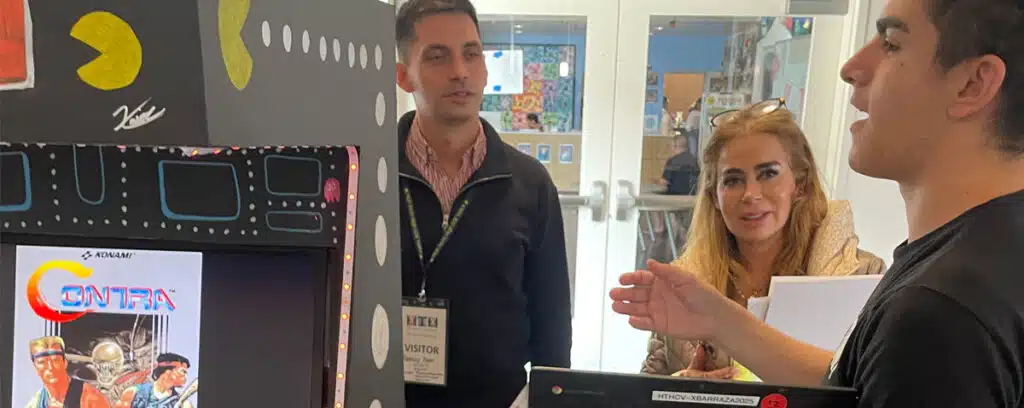 A young man explains an arcade game display to two visitors during one of the Curated Visits. The display features Pac-Man graphics and a screen showing the game Contra, capturing the guests attentive interest.