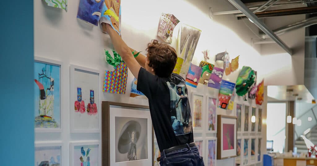 A person stands on tiptoe, reaching up to adjust colorful artwork as part of a thoughtful curation on a gallery-style wall lined with various framed and unframed art pieces in a well-lit, modern space.