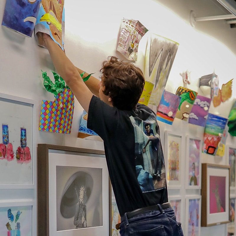 A person with short brown hair reaches up to adjust colorful, abstract artwork as part of a photo essay on creative curation, surrounded by framed pictures and vibrant paper art while wearing a black t-shirt and blue jeans.