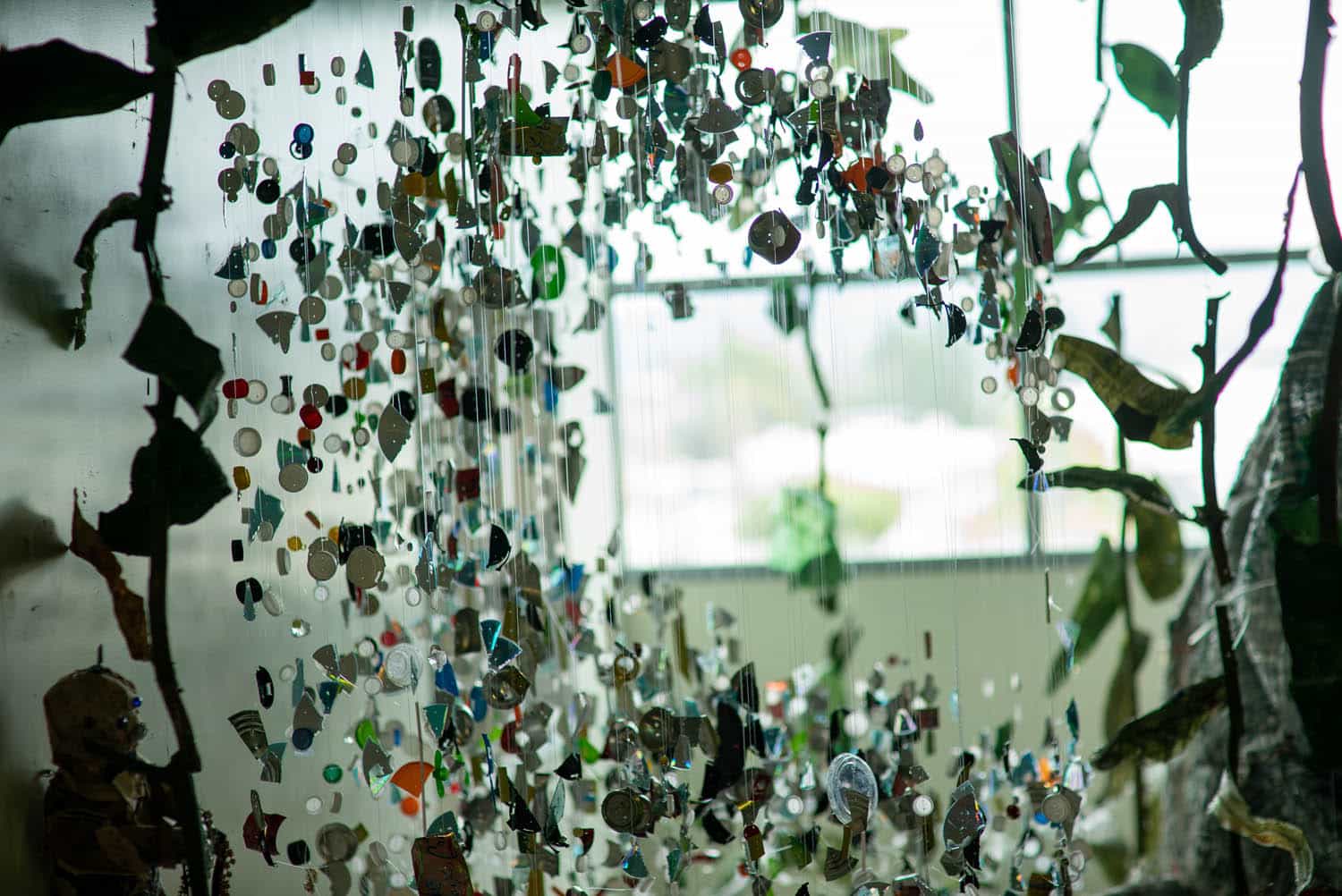 Colorful pieces of plastic and other materials hang from strings in a curated art installation, with window light shining through in the background and leafy decor framing the photo essay scene.