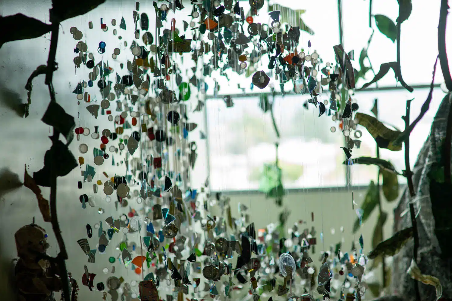 Colorful pieces of plastic and other materials hang from strings in a curated art installation, with window light shining through in the background and leafy decor framing the photo essay scene.