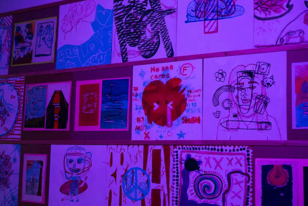 A wall covered with colorful abstract and cartoon-style drawings—including hearts, faces, and various shapes—glows under purple lighting. The artwork feels like a photo essay, with phrases like "No one cares" curating the mood.