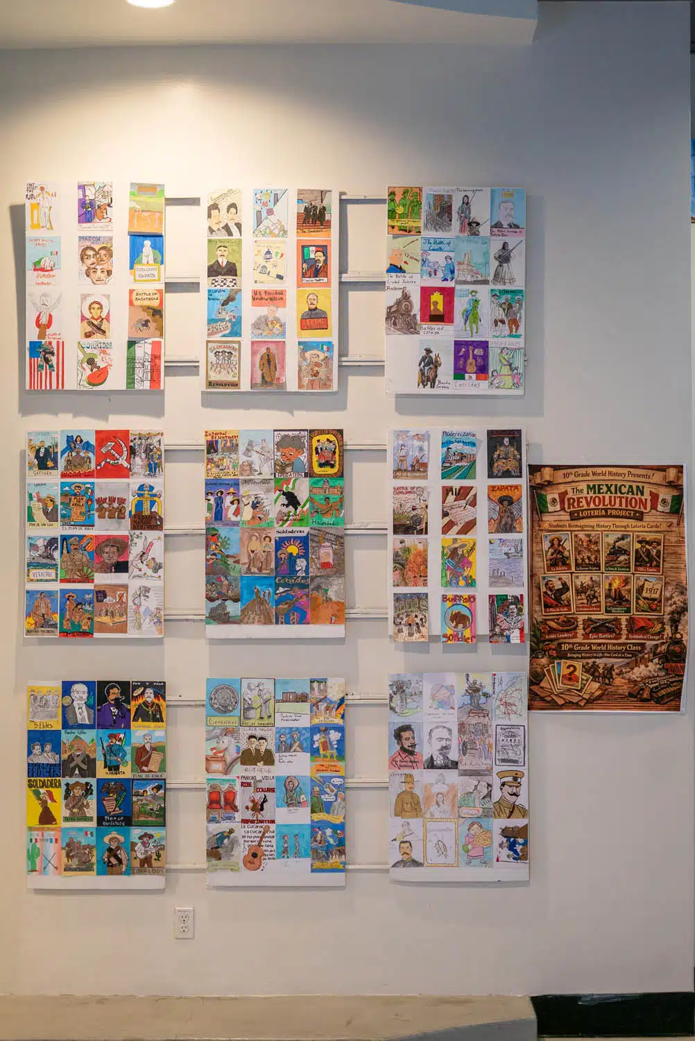 A wall display showcases a curation of colorful illustrated cards or small posters in neat rows and columns. To the right, a vintage-style Mexican Million Dollar poster adds flair. The well-lit indoor space feels like a living photo essay.