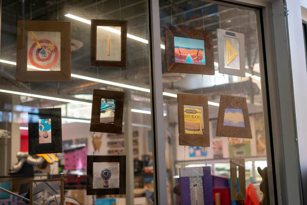 A glass window displays a curated selection of small, colorful art pieces in wooden frames, hanging in rows. The background reveals the interior of a creative, well-lit space filled with art supplies—perfect for a photo essay on artistic curation.