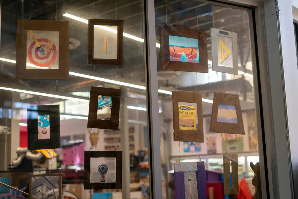 A glass window displays a curated selection of small, colorful art pieces in wooden frames, hanging in rows. The background reveals the interior of a creative, well-lit space filled with art supplies—perfect for a photo essay on artistic curation.