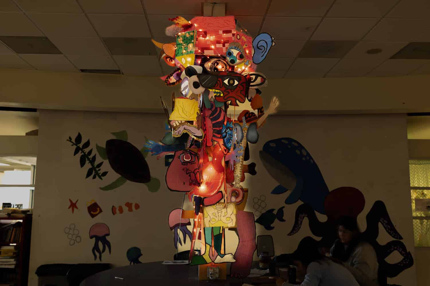 A tall, colorful sculpture—curated with cartoon faces, masks, and lights—stands on a table in a dimly lit room adorned with sea creature art. A person sits nearby at the table, contributing to a lively photo essay atmosphere.