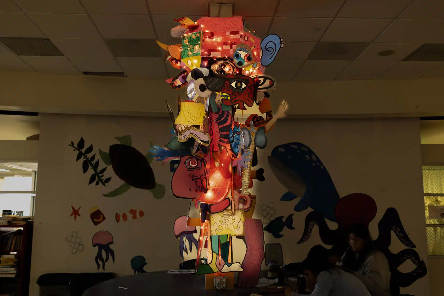 A tall, colorful sculpture—curated with cartoon faces, masks, and lights—stands on a table in a dimly lit room adorned with sea creature art. A person sits nearby at the table, contributing to a lively photo essay atmosphere.