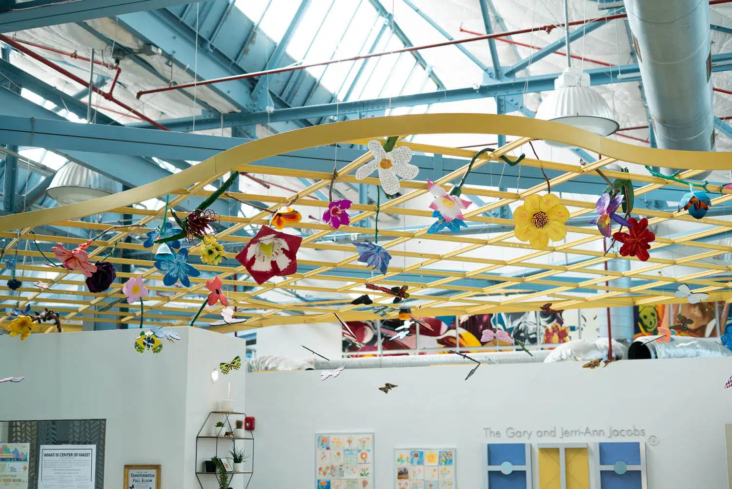 Colorful paper flowers and butterflies hang from a yellow grid structure on the ceiling of a bright room with skylights and blue beams—a playful curation, perfect for a photo essay, with art and children’s drawings decorating the white walls below.