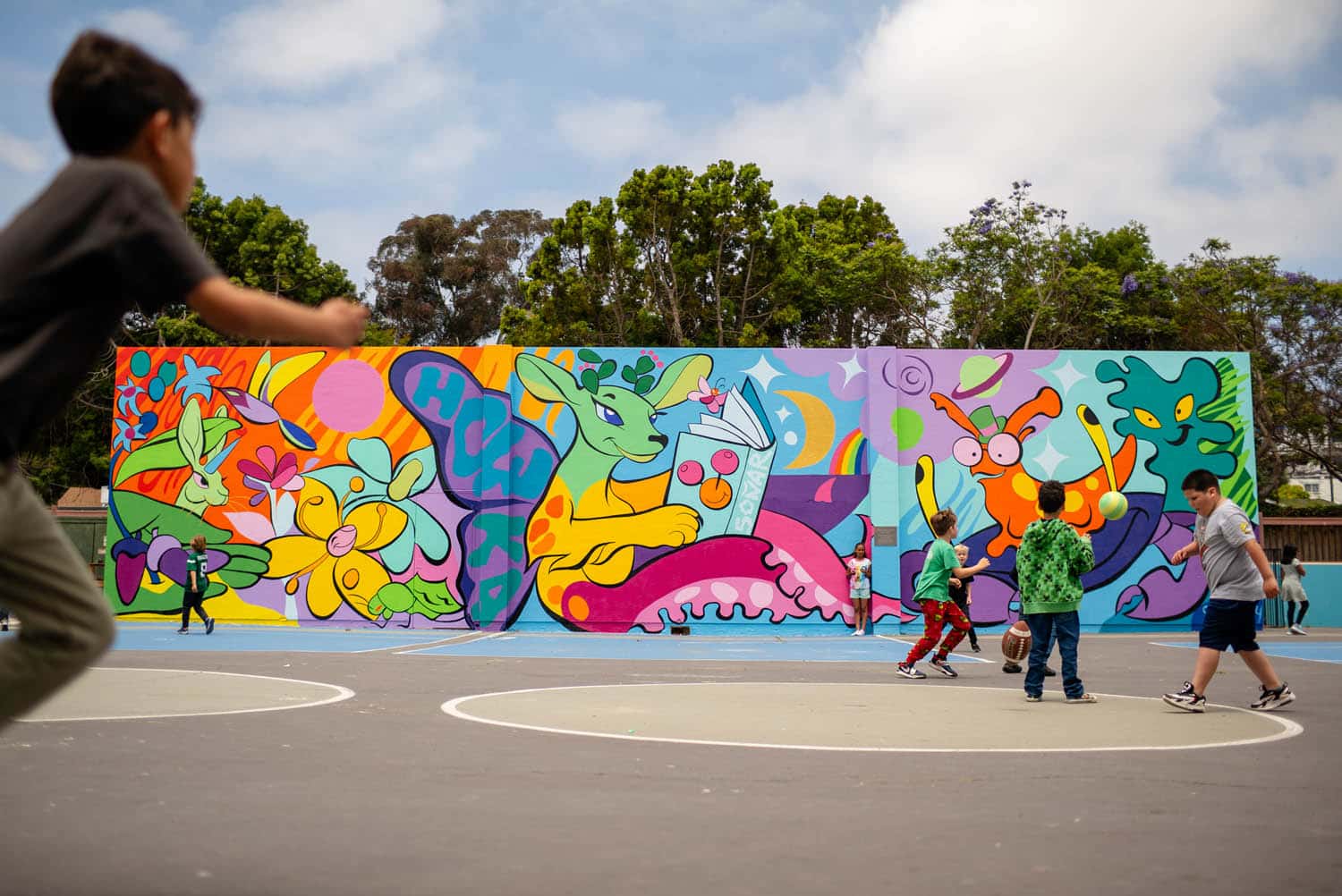 Children play basketball on an outdoor court in front of a colorful mural, creating a vibrant scene perfect for a photo essay curation that showcases whimsical animals, plants, and abstract shapes under a partly cloudy sky.