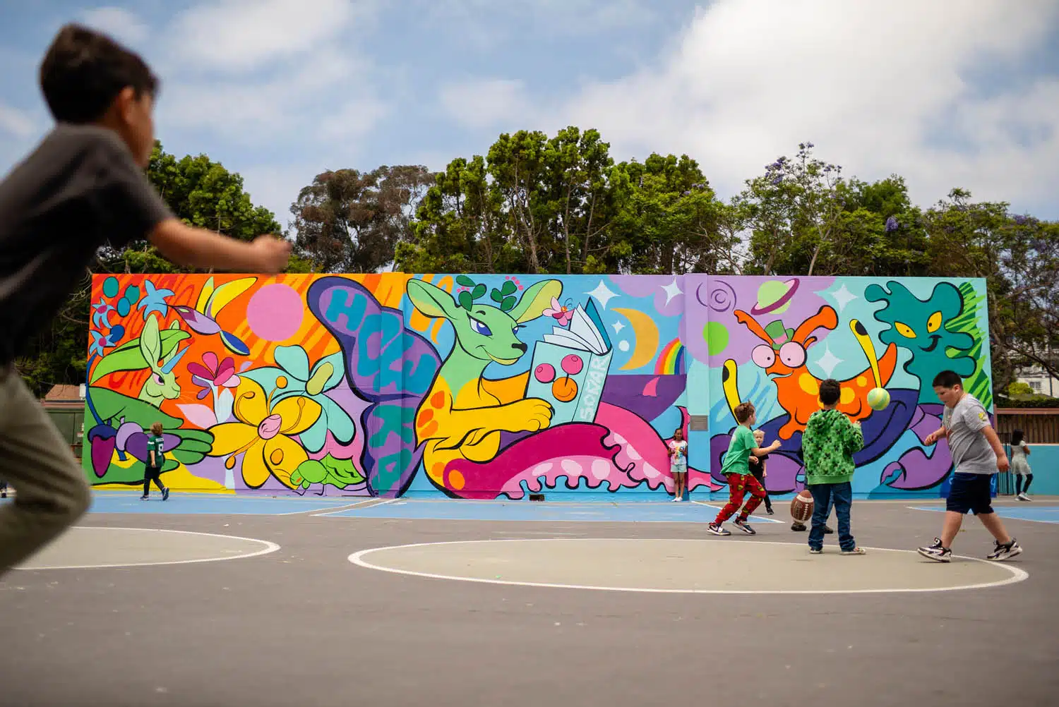 Children play basketball on an outdoor court in front of a colorful mural, creating a vibrant scene perfect for a photo essay curation that showcases whimsical animals, plants, and abstract shapes under a partly cloudy sky.