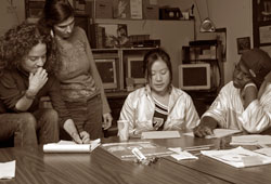 A group of four people in a room collaborate around a table, discussing and taking notes. There are computers and shelves in the background, conveying an office or study environment. The image is in sepia tone.