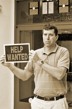 A man stands in front of a building holding a Help Wanted sign. The building has a sign above the door reading High Tech High. The man is wearing a short-sleeved polo shirt.
