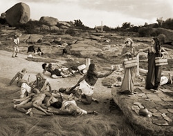 A sepia-toned image showing people in ancient-style attire, some lying on the ground and others standing. Two figures are exchanging items on a rocky terrain, with boulders and sparse vegetation in the background.