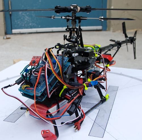 A remote-controlled helicopter sits on a sheet of paper. It is equipped with various electronic components and colorful wires attached to its frame, indicating modifications or customizations. The background is a blurred, blue-colored surface.