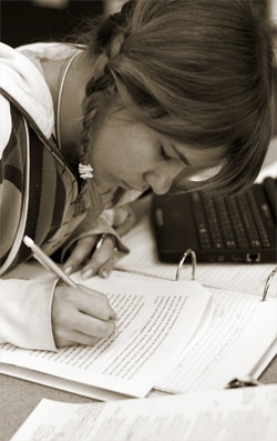 A person with braided hair is focused on writing in a notebook. A laptop and papers are visible on the desk. The image is in sepia tone, suggesting a thoughtful or studious atmosphere.