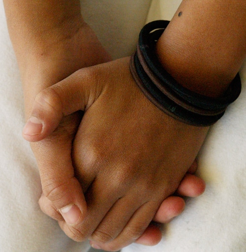 Two hands are clasped together. One wrist is adorned with multiple dark bracelets. The background is soft and neutral, possibly a blanket or fabric.