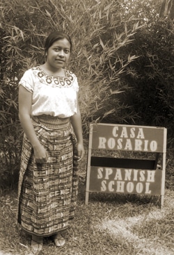 A woman stands beside a sign that reads Casa Rosario Spanish School in a grassy area with bamboo plants in the background. She is wearing a traditional embroidered top and patterned skirt. The photo is in sepia tones.