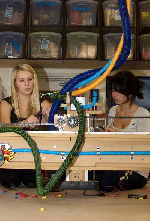 Two people working on a robotic project with colorful tubes and components, surrounded by storage bins. They are focused on assembling parts at a workspace.