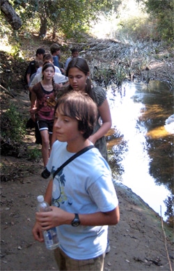 A group of people, including children and teenagers, walk along a wooded trail beside a stream. The scene is sunny, and the area is surrounded by lush greenery. One person in the foreground holds a water bottle.