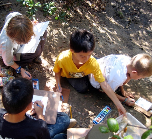 Four children sit on the ground outdoors, painting with watercolors. They are focused on their work, surrounded by natural light and greenery. Art supplies are scattered around them as they use clipboards to support their paper.