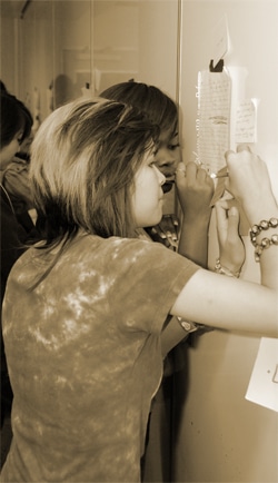 A person with two-toned hair writes a message on a piece of paper attached to a wall, surrounded by other papers. Another person is visible in the background. The image is in sepia tones.