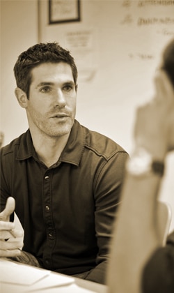 A man with short dark hair is sitting at a table, speaking and gesturing with his hands. He is wearing a dark button-up shirt. The setting appears to be a casual indoor environment, with another person partially visible in the foreground.