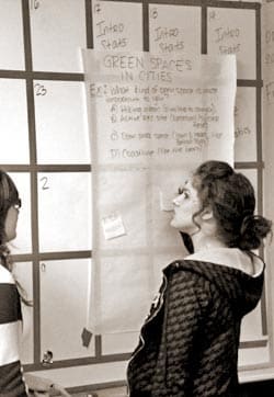 Two people stand in front of a grid-covered wall with notes. A sheet with the heading Green Spaces in Cities is pinned on the wall. Theyre engaged in discussing or reviewing its content. The image is in black and white.