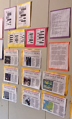 A wall display features several editions of The Daily News newspaper, each bordered with different colored paper. Above are printed documents with charts and text, bordered in various colors, organized neatly in rows.