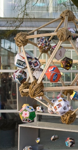 A geometric sculpture made of wooden rods and twine forms a large polyhedron. Various smaller polyhedra with colorful designs are attached, resembling a molecular structure, set against a glass building backdrop.