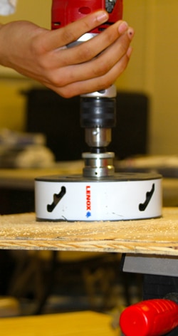 Close-up of a persons hand using a LENOX hole saw attached to a drill to cut through a wooden surface. Wood shavings are visible on the surface. The background is out of focus, emphasizing the tool and hands.