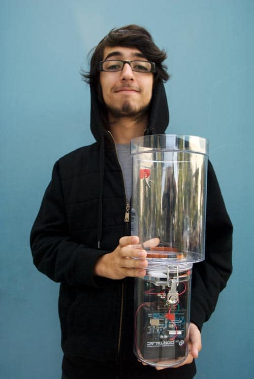 A person wearing a black hoodie, standing against a blue background, holds a large transparent cylindrical container. Inside the container are electronic components and wiring, visible through the clear surface.
