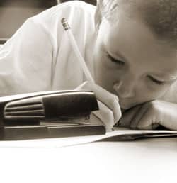 A child is intently writing on a piece of paper with a pencil, resting their head on their arm. A stapler is visible on the table next to them. The image is in black and white.