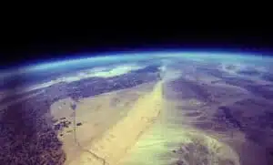 View of Earth from space showing a vast desert landscape with sand dunes and rocky terrain, under a curved horizon. The atmosphere creates a blue halo at the edge of the planet against the darkness of space.