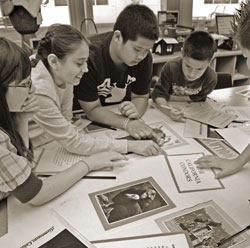 A group of students is gathered around a table, engaged in a collaborative activity. They are examining papers and images spread out before them, appearing focused and engaged. The setting looks like a classroom.