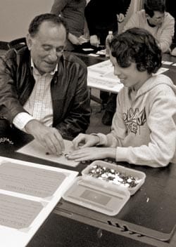 A man and a young person are engaged in a hands-on activity at a table, surrounded by containers of small items. The man is assisting the young person with organizing or assembling objects. They are in a classroom or workshop setting.