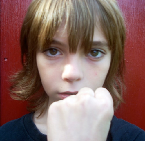 A child with shoulder-length brown hair is looking directly at the camera, holding their fist up in front. The background is a solid red color.
