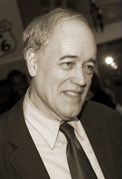 A man in a suit and tie is smiling in a candid photograph. The background is blurred, featuring indistinct shapes and a hint of signage. The image has a sepia tone.
