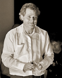 A man wearing a white long-sleeve shirt, standing and holding something in his hand. The image is in sepia tone, and he seems to be speaking or performing in front of an audience.
