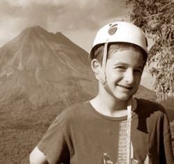 A young person wearing a climbing helmet smiles at the camera with a volcano in the background. The image is in sepia tones, and there is foliage on the right side.