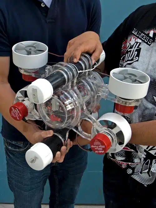 Two people holding a transparent underwater drone with multiple propellers on its sides. The drone has visible electronic components and red end caps. They are standing against a blue background.