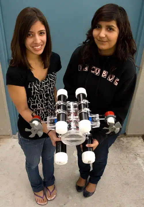 Two people standing and holding a small underwater robot with propellers. They are smiling and posing on a concrete surface with a blue door in the background.