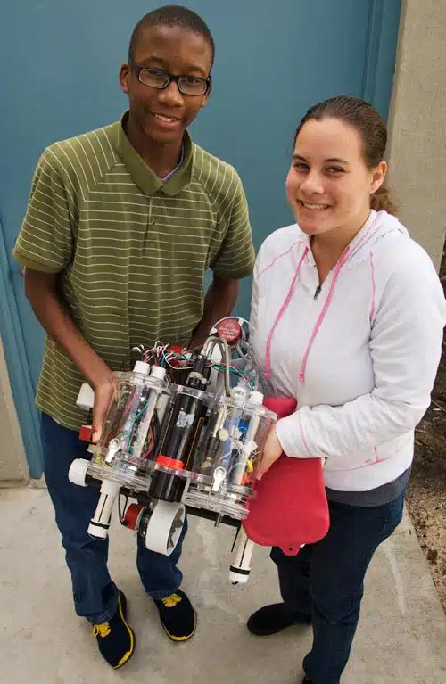 Two people smile while holding a small robot with wheels and transparent cylinders. The person on the left wears glasses and a green striped shirt. The person on the right wears a white and pink hoodie. They stand in front of a blue wall.