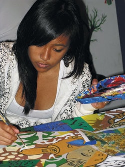 A person is focused on painting colorful designs on a flat surface. They hold a palette in one hand and a brush in the other, wearing a white patterned jacket. The scene appears artistic and creative.