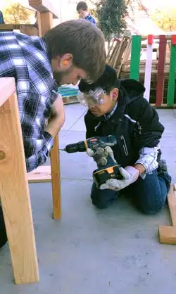 An adult and a child are assembling a wooden structure outdoors. The child wears safety goggles and ear protection while using a power drill. The adult leans in to assist. Various wooden planks and tools are scattered around them.