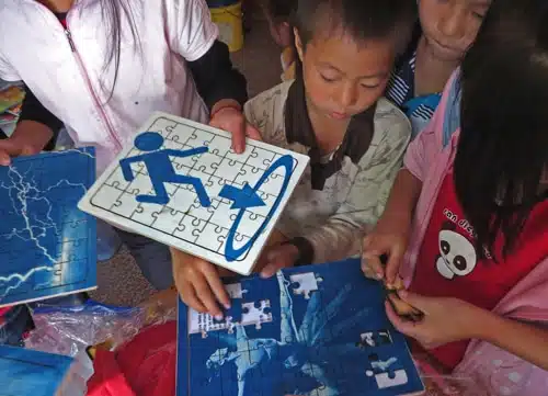 Children working on puzzle pieces with blue and white designs. One holds a completed puzzle with stick figures and arrows, while others are assembling a puzzle with a different blue image. They are gathered closely and focused on the activity.
