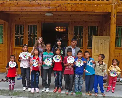 A group of children and a few adults standing in front of a wooden building, holding decorated paper plates. The children are smiling, and the adults are standing behind them. The group is casually dressed.