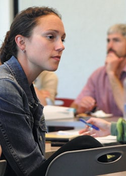 A woman with long brown hair, wearing a denim jacket, is attentively listening in a classroom setting. She sits at a table with notebooks and pens, and a man in a pink shirt is seated in the background, slightly out of focus.